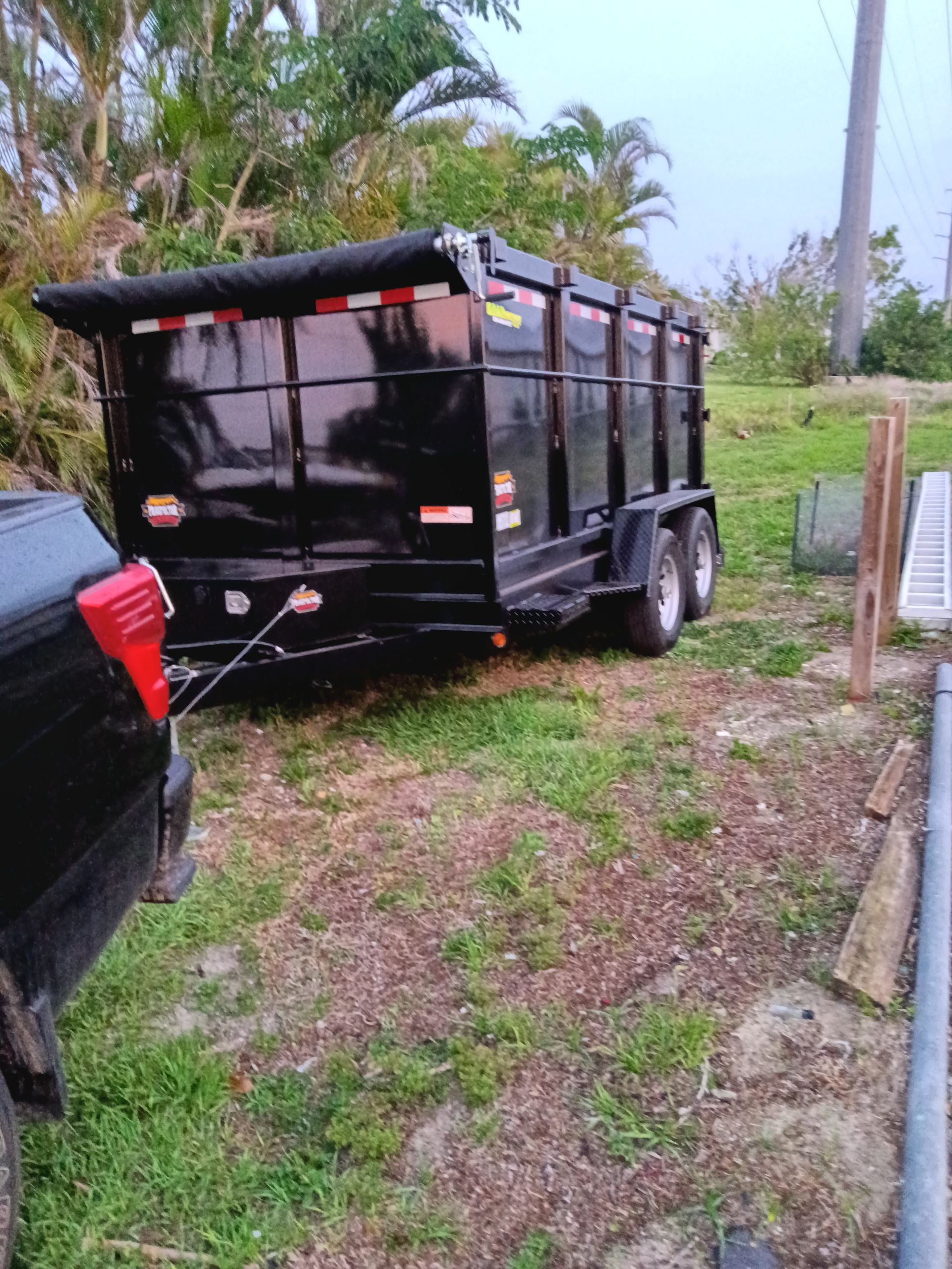 Black dump trailer hitched to a black truck, parked on grass next to a wooden fence.