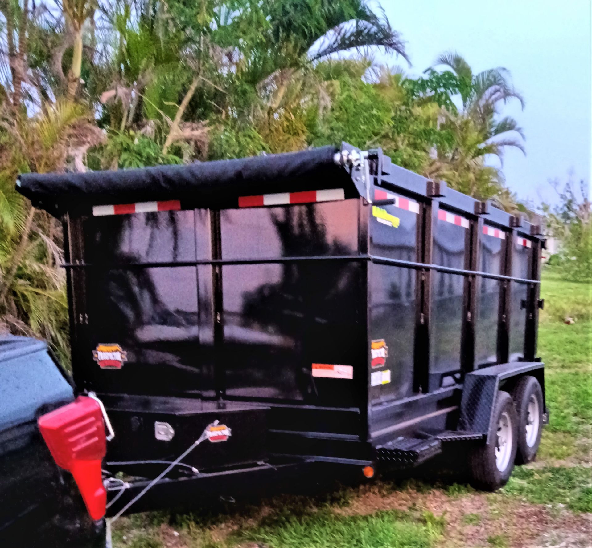 Black dump trailer hitched to a vehicle on a grassy area, trees in the background.