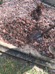 Shovel digging in a bed of reddish rocks and soil beside a concrete edge and grass.