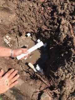 Person holding white PVC pipe in a muddy hole, possibly fixing a broken pipe in a yard.