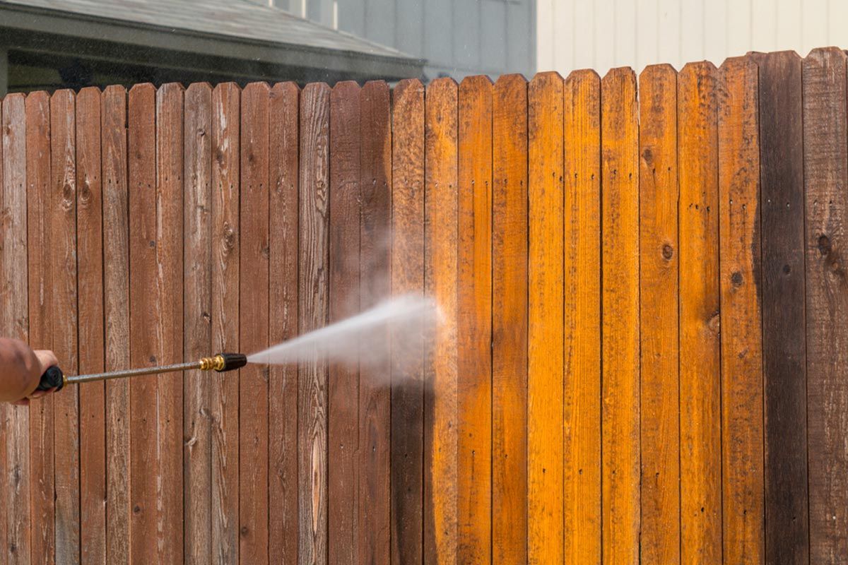 A person is using a high pressure washer to clean a wooden fence.
