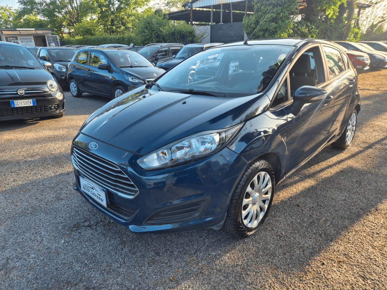 Una Ford Fiesta blu scuro parcheggiata in un parcheggio insieme ad altre auto in una giornata di sole.