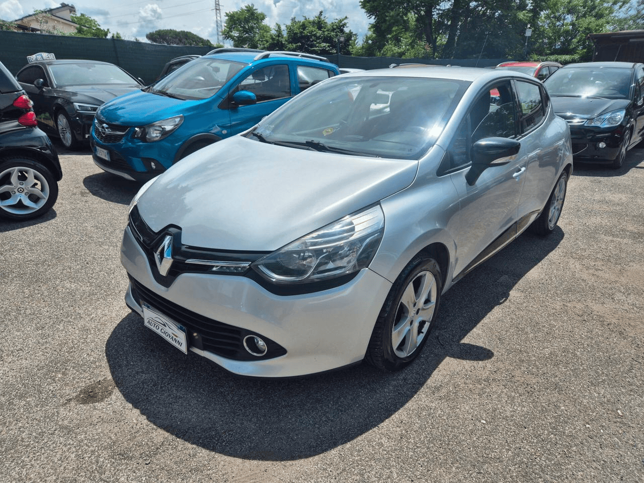 Una Renault Clio argentata parcheggiata in un parcheggio insieme ad altre auto in una giornata di sole.