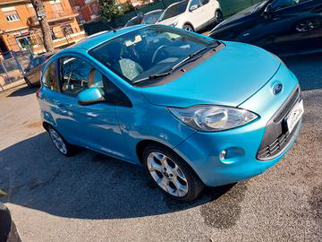 Una piccola Ford Ka blu brillante parcheggiata sull'asfalto. L'auto ha linee arrotondate e cerchi argentati.