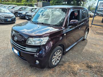 Un'auto Toyota bB color marrone scuro parcheggiata all'aperto su un terreno ghiaioso, con altre auto visibili sullo sfondo.