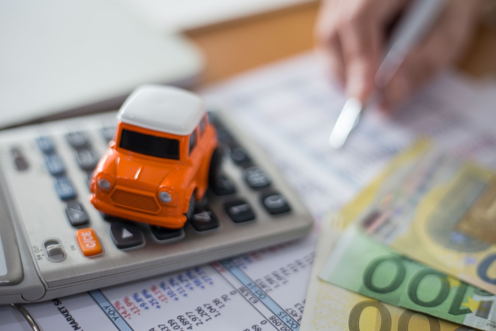 Una macchinina arancione su una calcolatrice con soldi e documenti, sullo sfondo qualcuno che scrive con una penna.