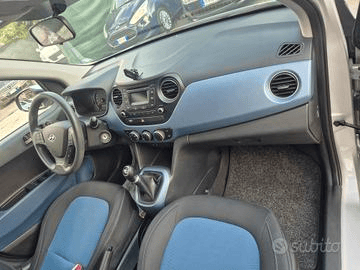 Vista interna di una Hyundai i10, che mostra il cruscotto, il volante, i sedili e la leva del cambio. Il cruscotto ha un inserto blu.