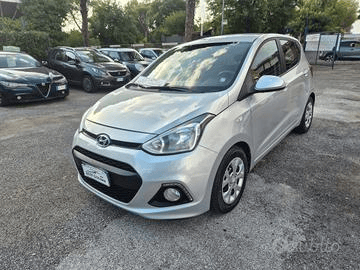 Hyundai i10 argentata parcheggiata su un piazzale ghiaioso, con altre auto sullo sfondo. Ripresa diurna, cielo nuvoloso.