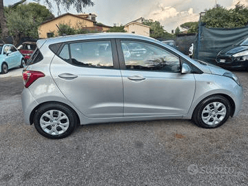 Hyundai i10 berlina color argento parcheggiata sulla ghiaia. Scatto diurno con altre auto ed edifici visibili sullo sfondo.