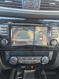 Cruscotto di un'auto con lo schermo di navigazione che mostra una visuale della telecamera posteriore e una vista dall'alto dell'auto, circondata da pulsanti e comandi del climatizzatore.