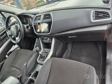 Vista interna del cruscotto di un'auto Suzuki con volante, console centrale e sedili. Sono visibili dettagli neri e argento.