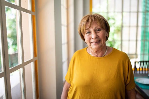 An elderly woman in a yellow shirt is standing in front of a window.