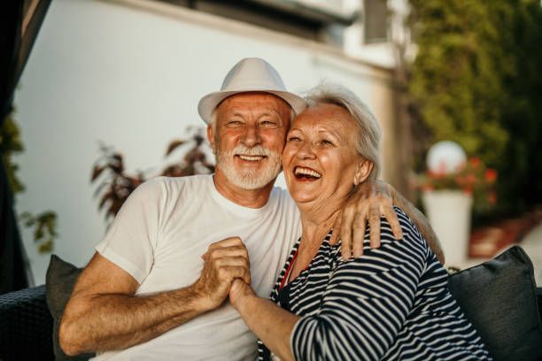 An elderly couple is sitting on a couch holding hands and smiling.