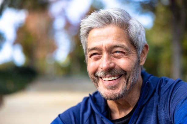 A man with gray hair and a beard is smiling in a park.
