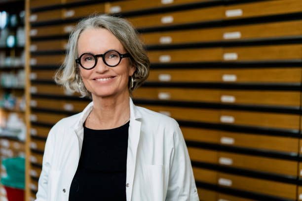 A woman in a lab coat and glasses is smiling in a pharmacy.