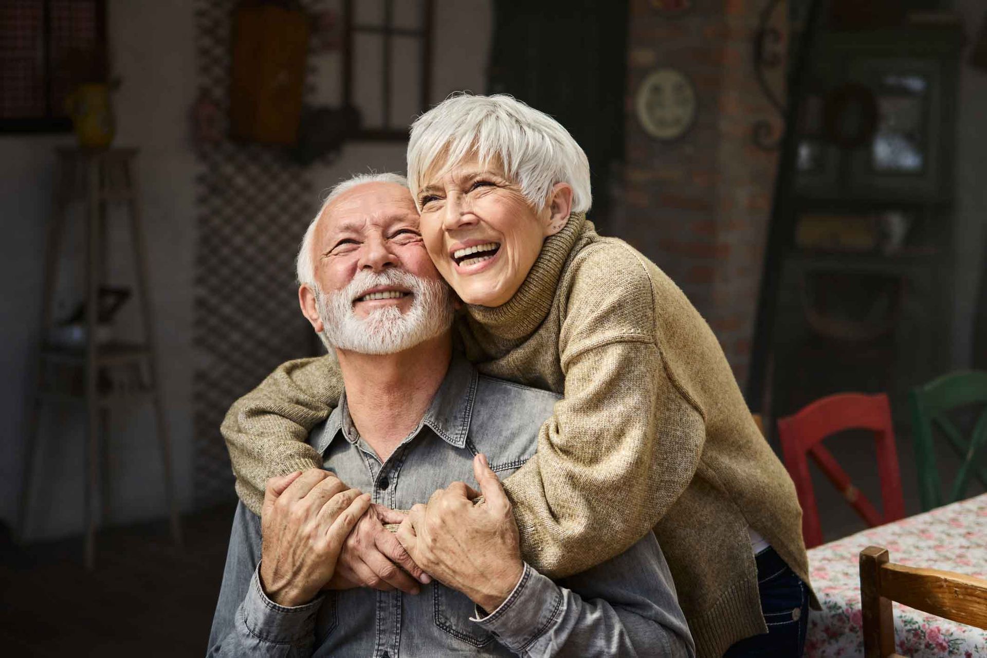 An elderly couple is hugging each other and smiling.