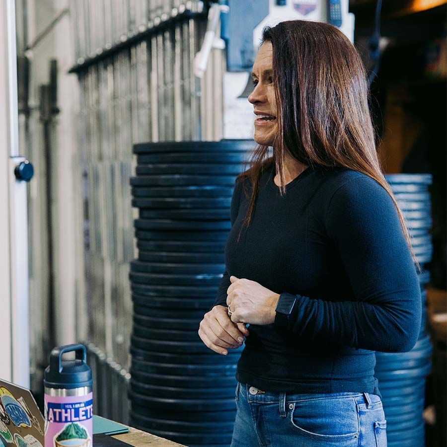 A person with long brown hair in a long-sleeved black shirt talks in a gym setting with stacked weight plates in the back.