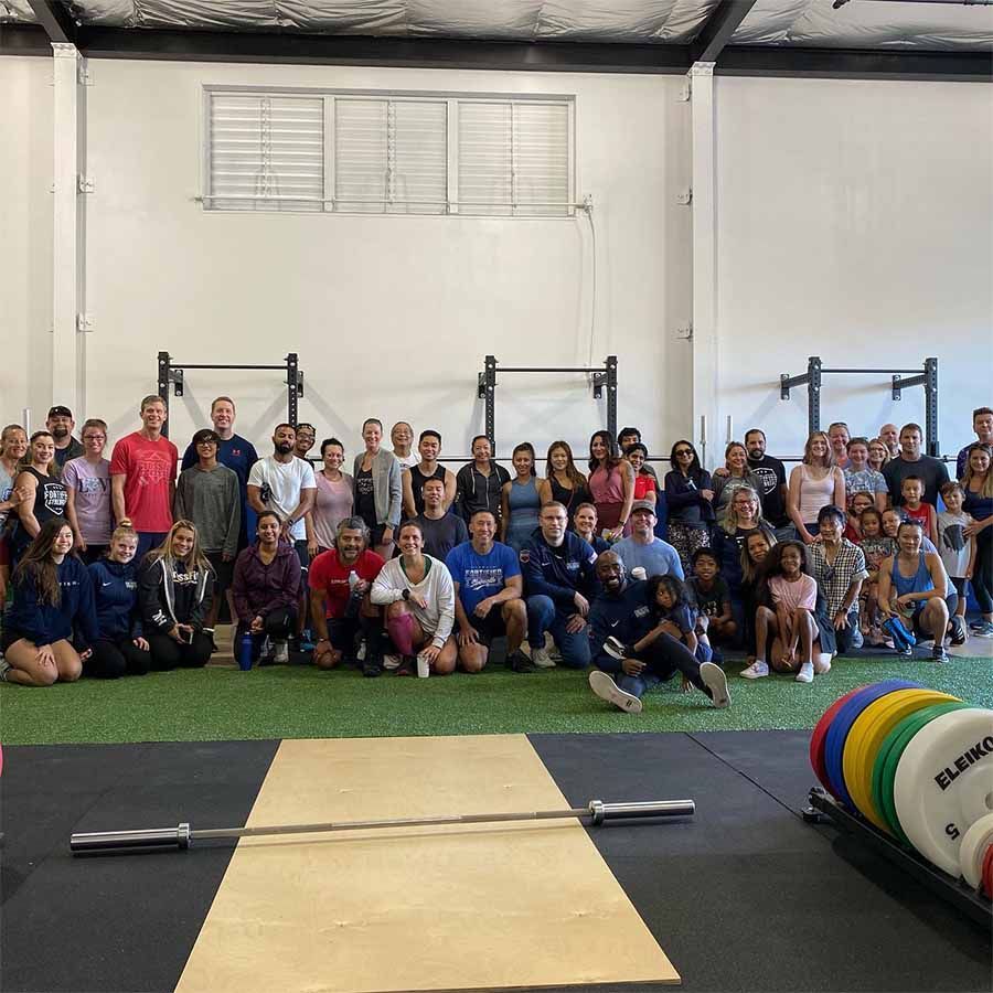 A large group of people smiling and posing in a gym with weightlifting equipment in the foreground.