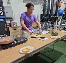 A person in a purple shirt slices greens on a wooden cutting board at a table set up for cooking in a gym.