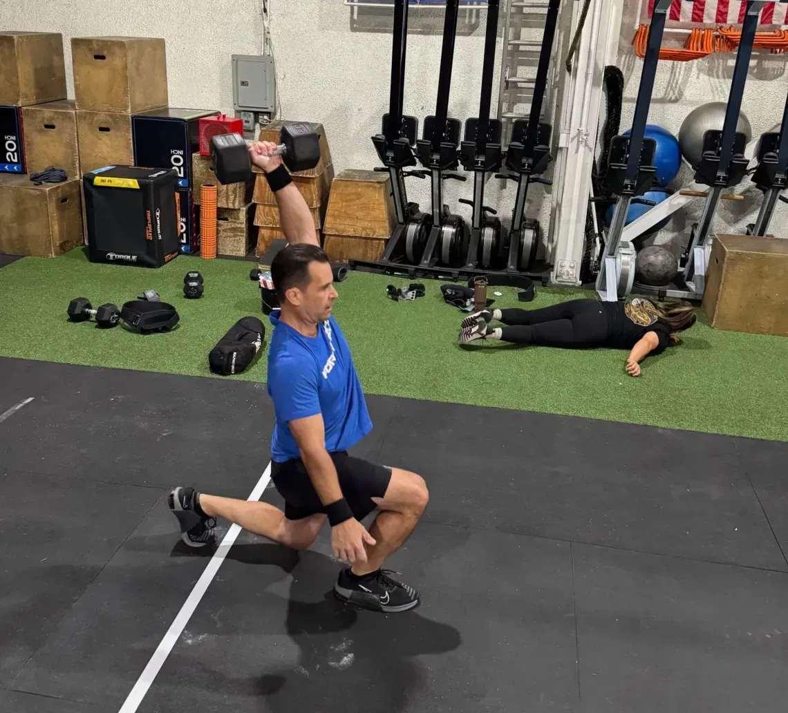 A person in a blue shirt performs a lunge while holding a dumbbell overhead in a gym, with another person resting nearby.