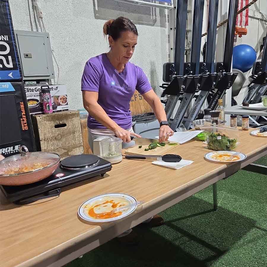 A person in a purple shirt slices greens on a wooden cutting board at a table set up for cooking in a gym.