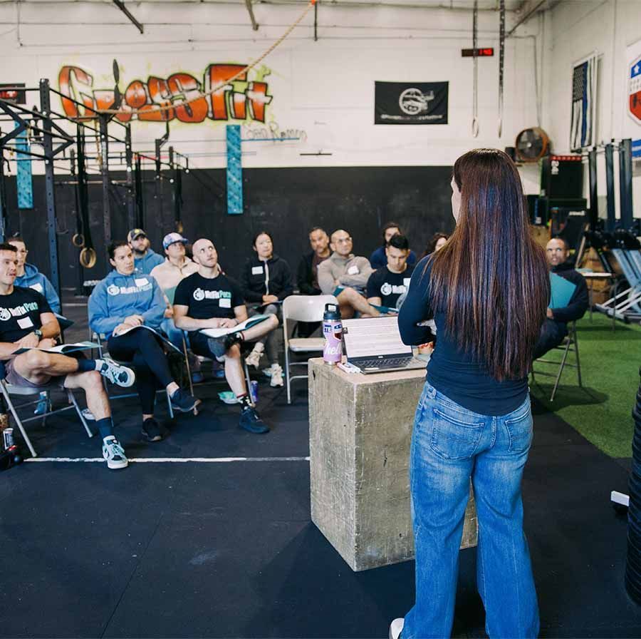 A group of people sit in chairs inside a gym, listening to a speaker standing at a wooden block podium with a laptop.