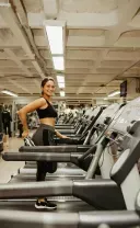 A person in athletic wear smiling while stretching on a treadmill in a brightly lit gym.
