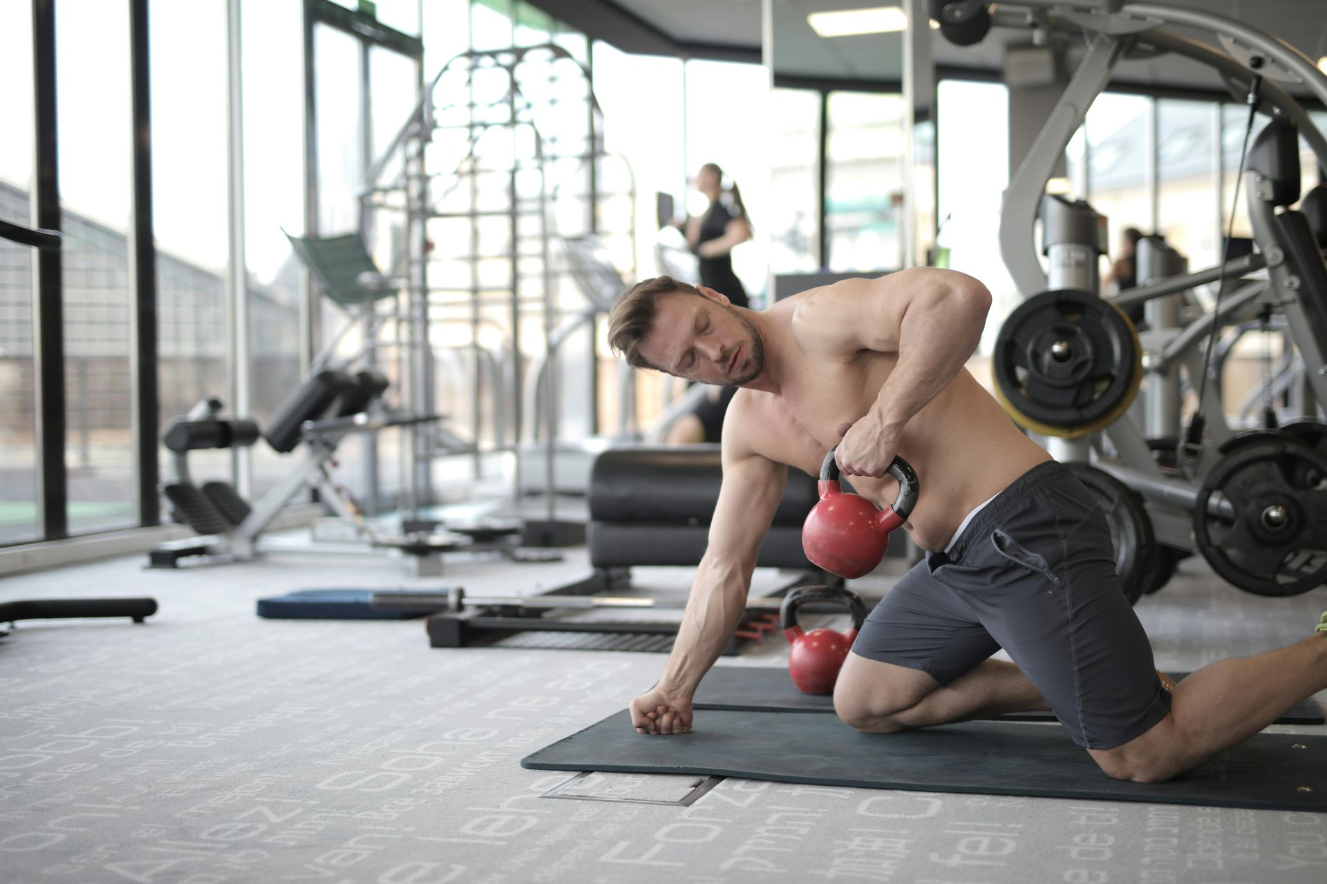 A shirtless person kneeling on a gym mat performs a one-arm row exercise with a red kettlebell in a gym setting.
