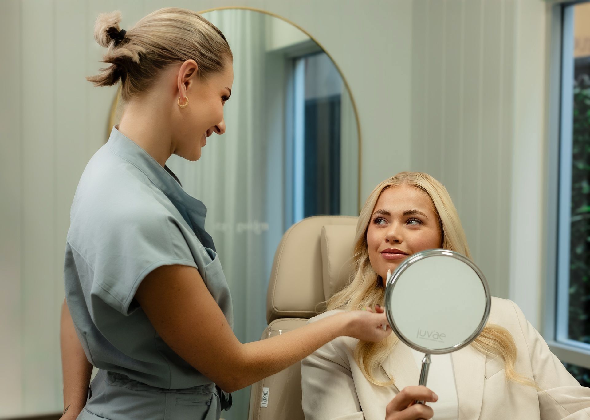 A woman examines her face in a mirror as a healthcare professional points to it in a medical office.