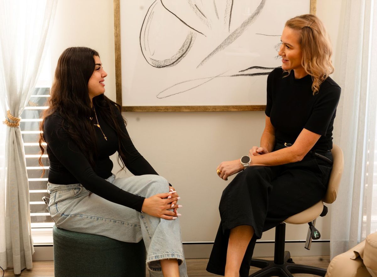 Two women in a well-lit room converse. One sits on a stool, the other on a chair. They smile and gesture.