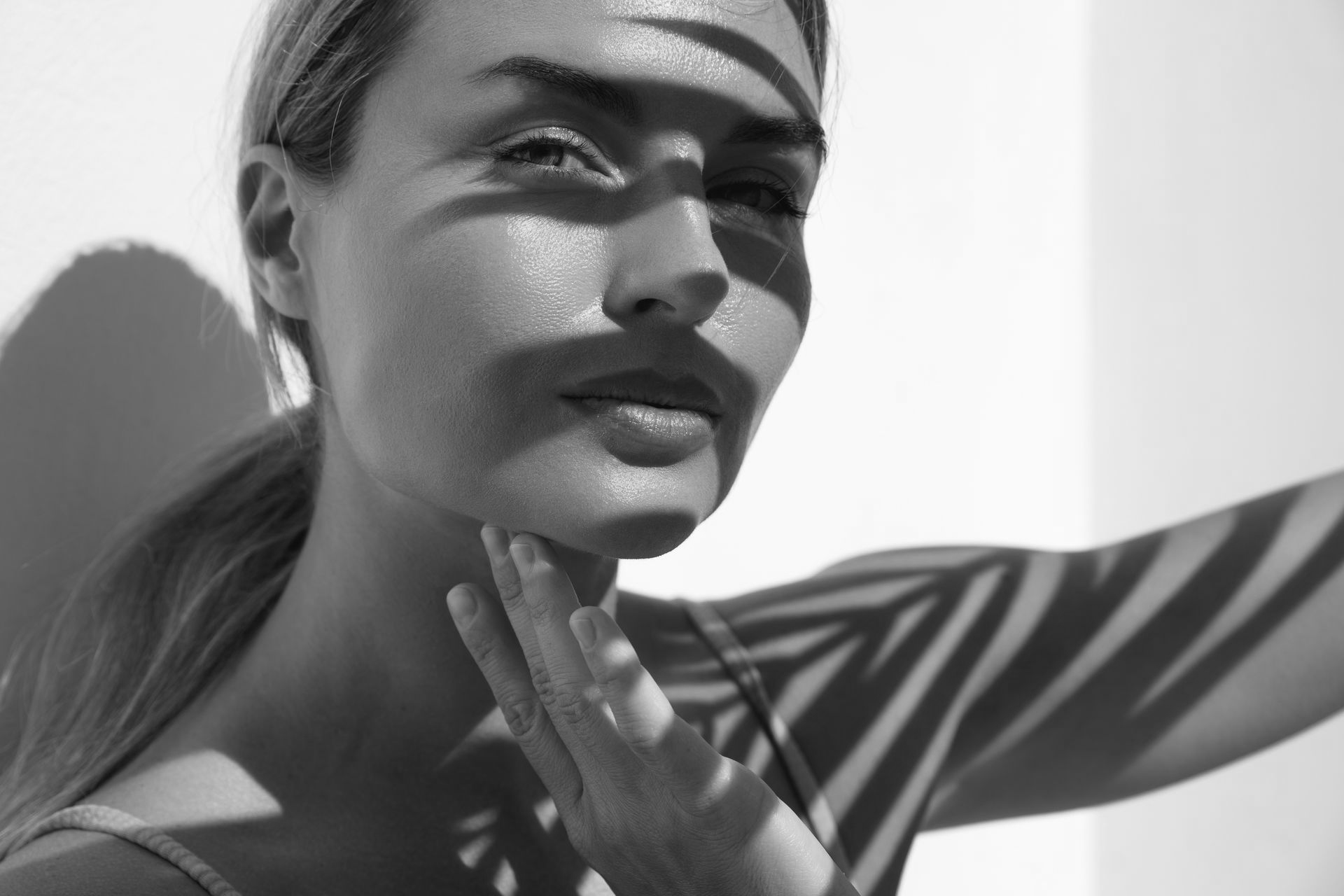 Woman's face with palm frond shadows, hand near chin. Black and white, sunlit.