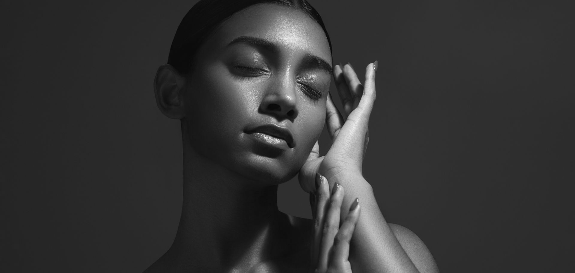 A woman with closed eyes in a black and white studio portrait, hands near her face, bathed in light.