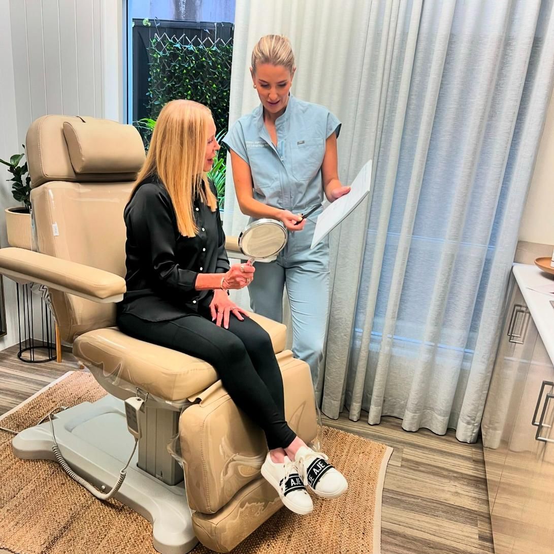 A blonde woman consults with a professional in a beige aesthetician's chair. They are in a bright room.