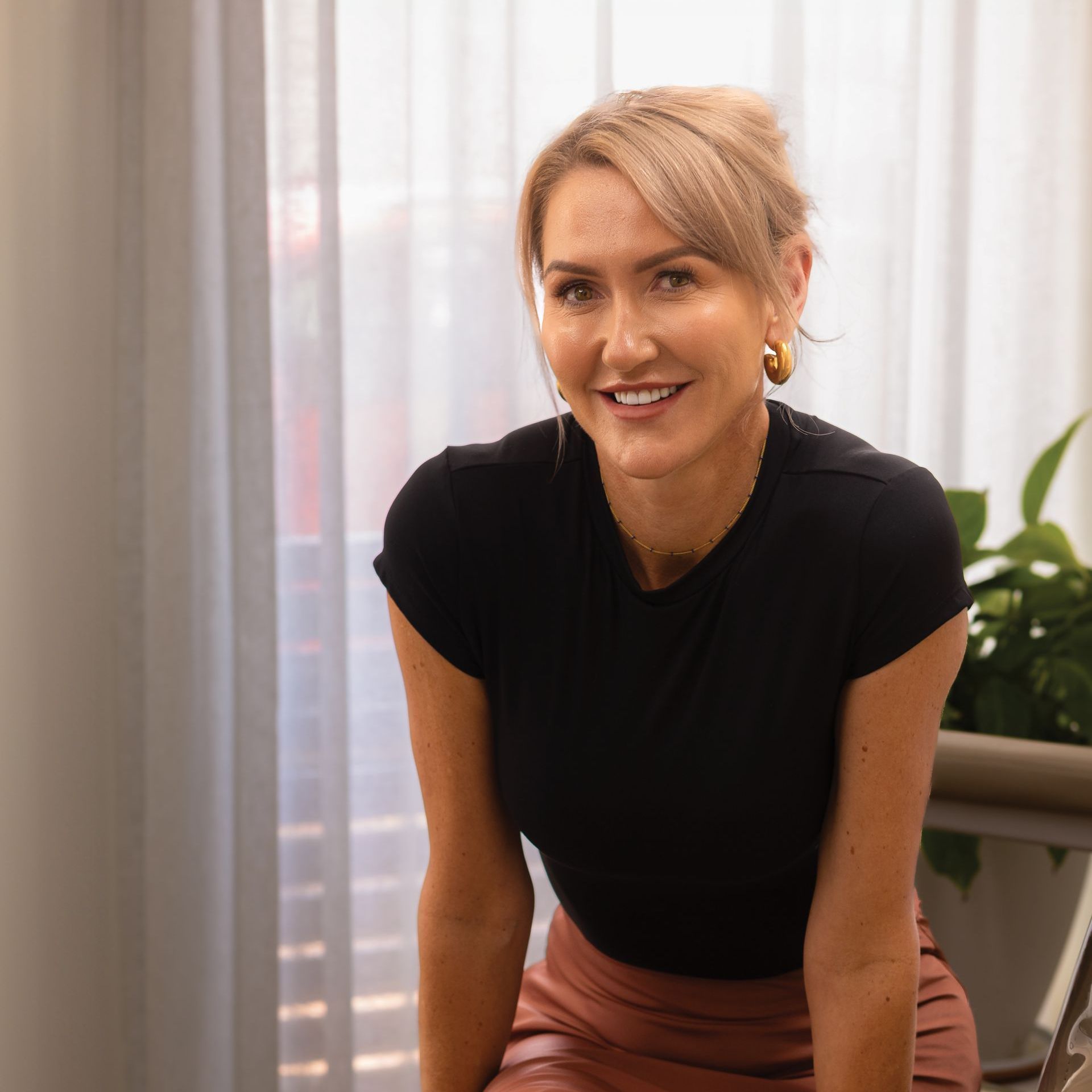 Woman with blonde hair smiles, wearing a black shirt and pink skirt, indoors by a window and a plant.