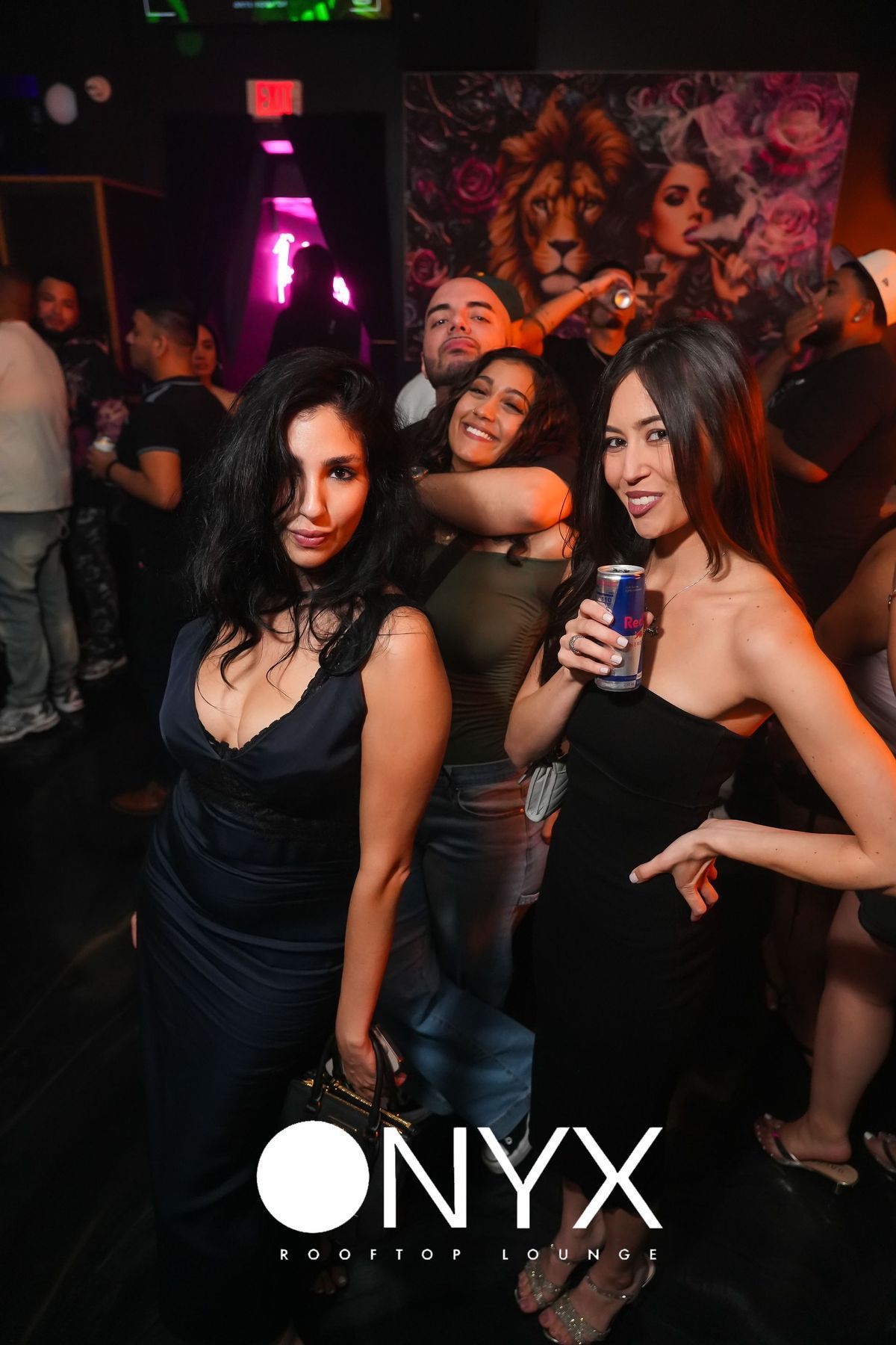 People at Onyx nightclub, woman in black dress holds Red Bull. Others are smiling, dark interior.