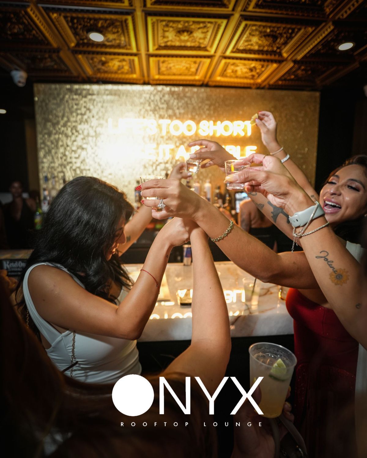 Women toasting shots at a bar, Onyx Soul Up Lounge logo in foreground. Golden ceiling, bright sign, and happy expressions.