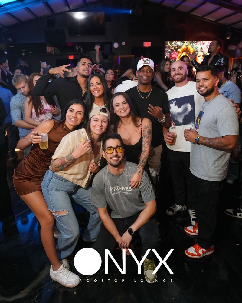 Group of people at Onyx club, posing for photo. People smiling, some holding drinks. Dark setting, logo in foreground.