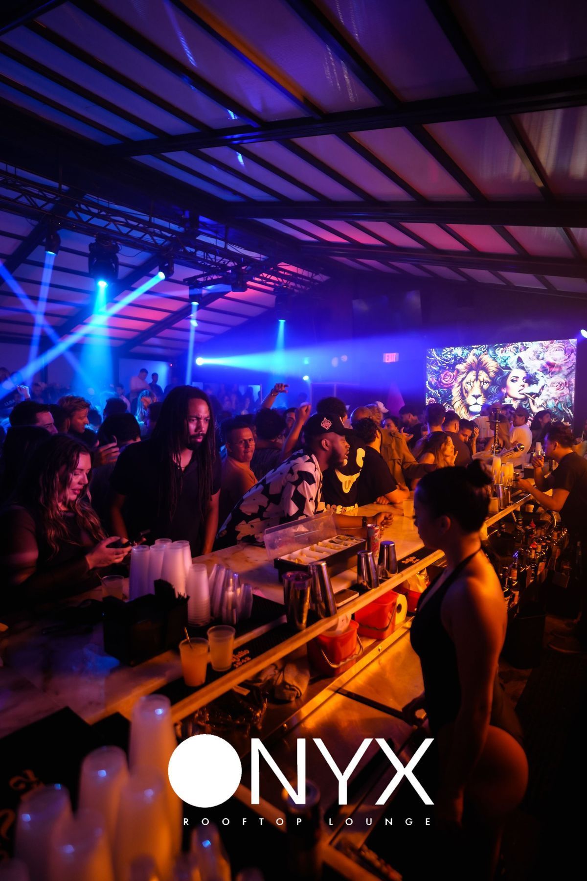 Nightclub interior with a bar and crowd. Blue, pink, and white lights illuminate the scene. 