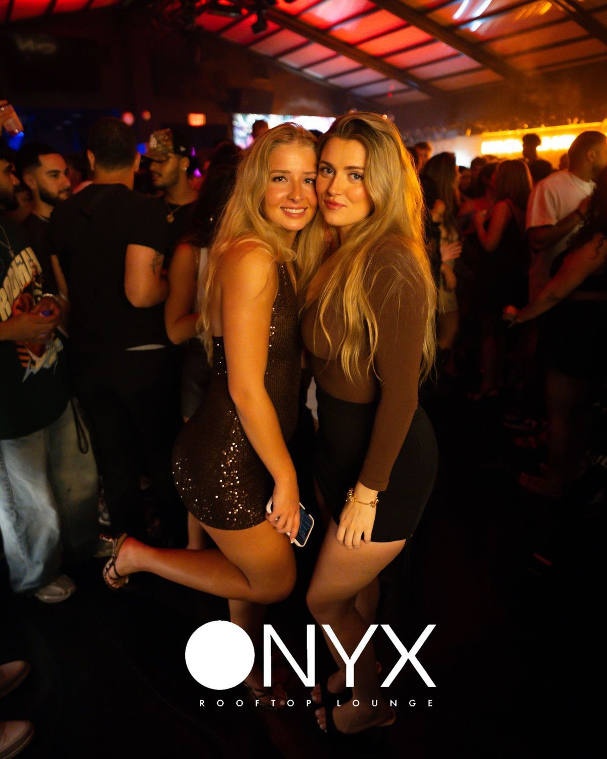 Two young women smiling at a night club, one in a brown dress, the other in a sparkly one. Club's name: ONYX.