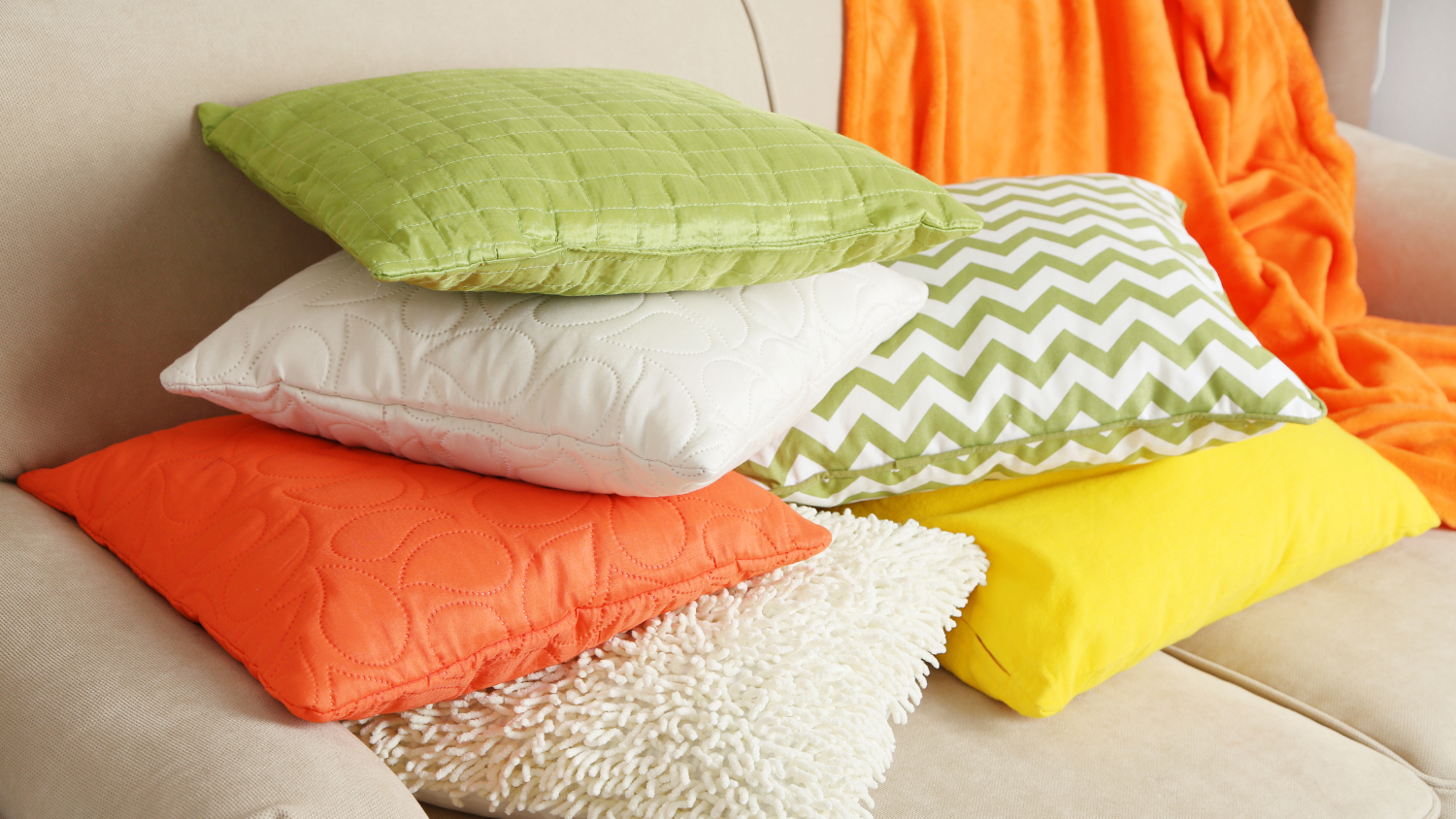 Stapel bunter Kissen auf einem beigen Sofa: grün, weiß, orange, gelb, mit einer orangefarbenen Decke im Hintergrund.
