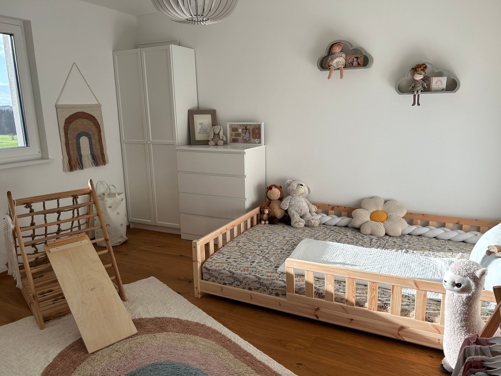 Kinderzimmer mit Holzbett, Spielstruktur und Regenbogenteppich. Weiße Kommode und Kleiderschrank.