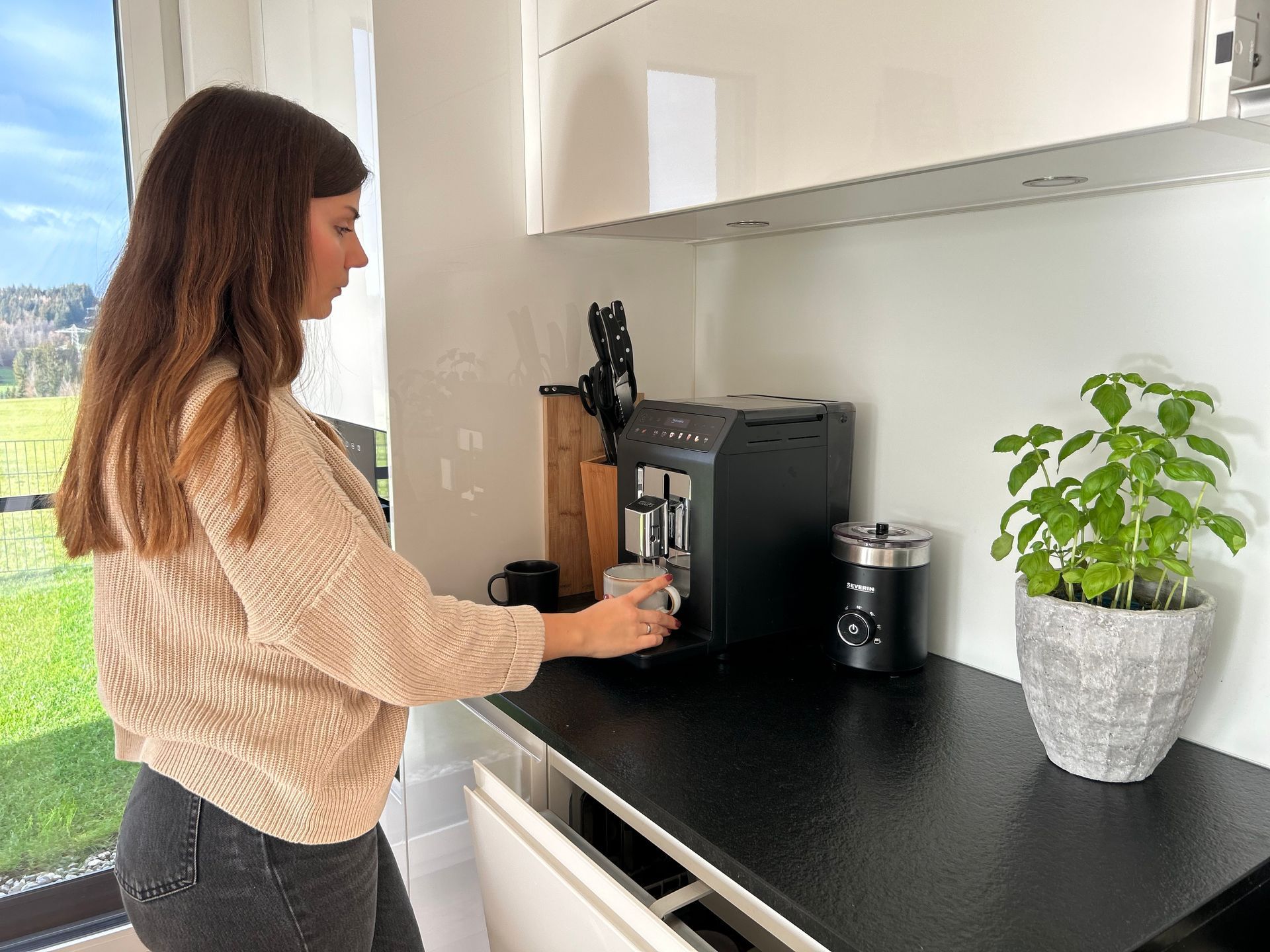Frau macht Kaffee in einer modernen Küche, greift nach einer Tasse neben einer schwarzen Espressomaschine, Blick aus dem Fenster.