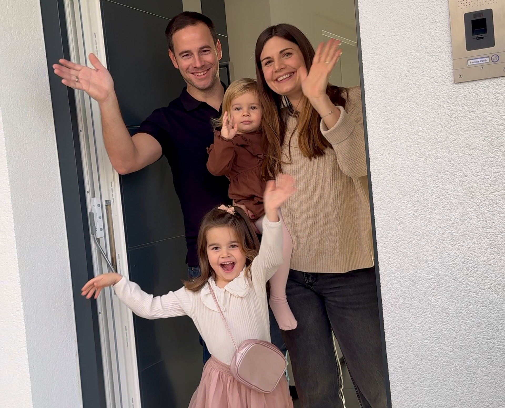 Eine vierköpfige Familie winkt aus einer offenen Tür: Eltern, zwei kleine Töchter.