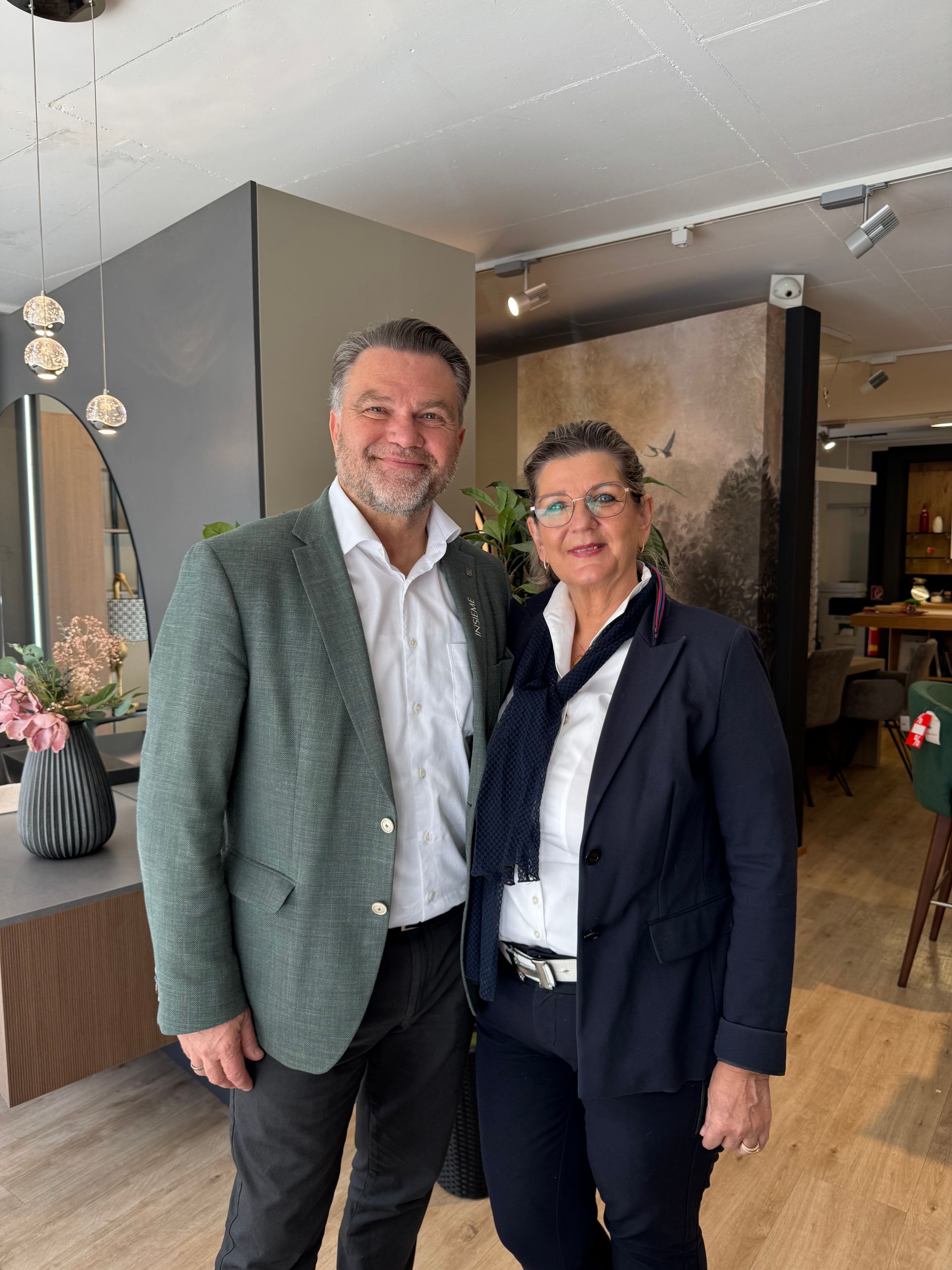 Mann und Frau stehen im Innenbereich; Mann in grüner Jacke, Frau im Blazer, beide lächeln.