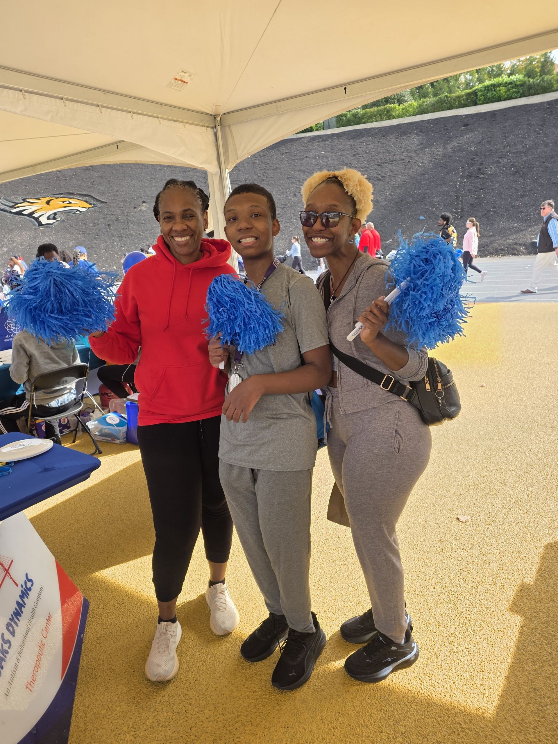 Three people smiling, holding blue pom-poms under a tent. 