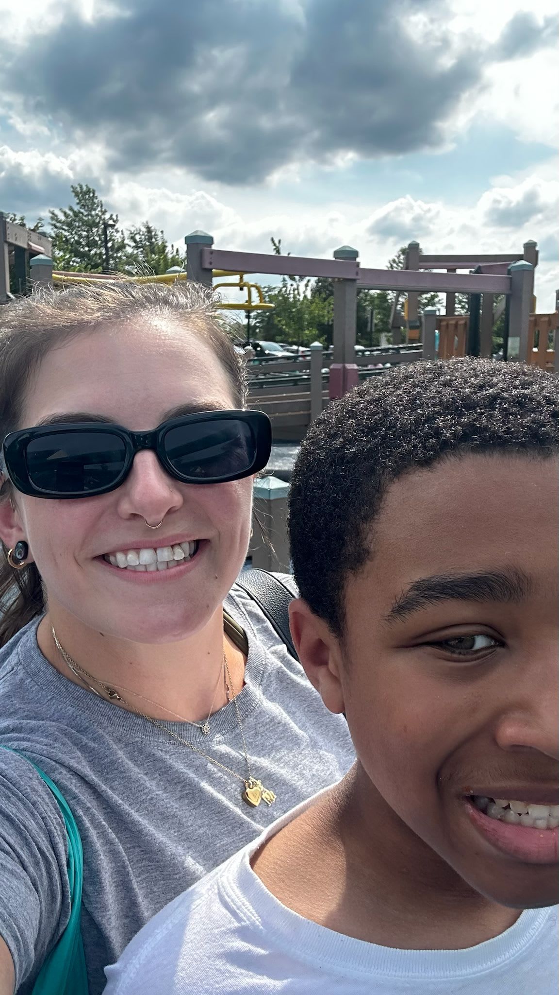 Woman in sunglasses smiles next to a boy with braces, both outdoors near a playground.