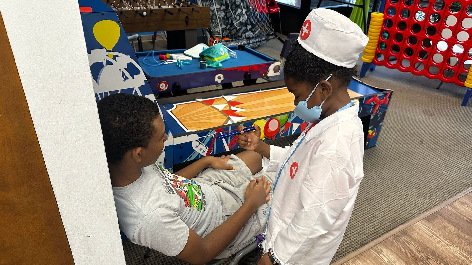 Child dressed as a doctor examines another person lying down, in a playroom with games.
