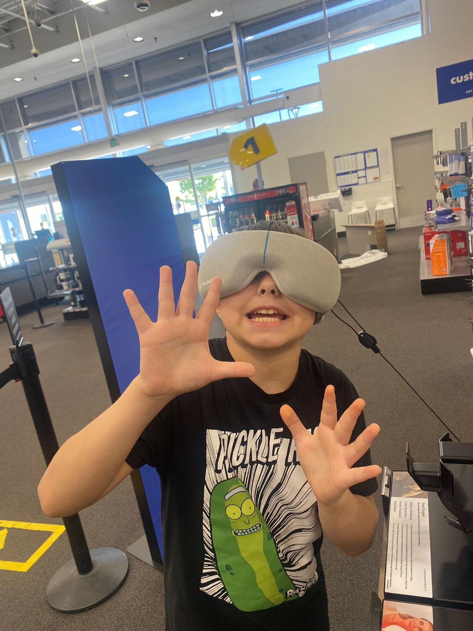 Boy wearing a gray sleep mask at a store, holding up his hands.