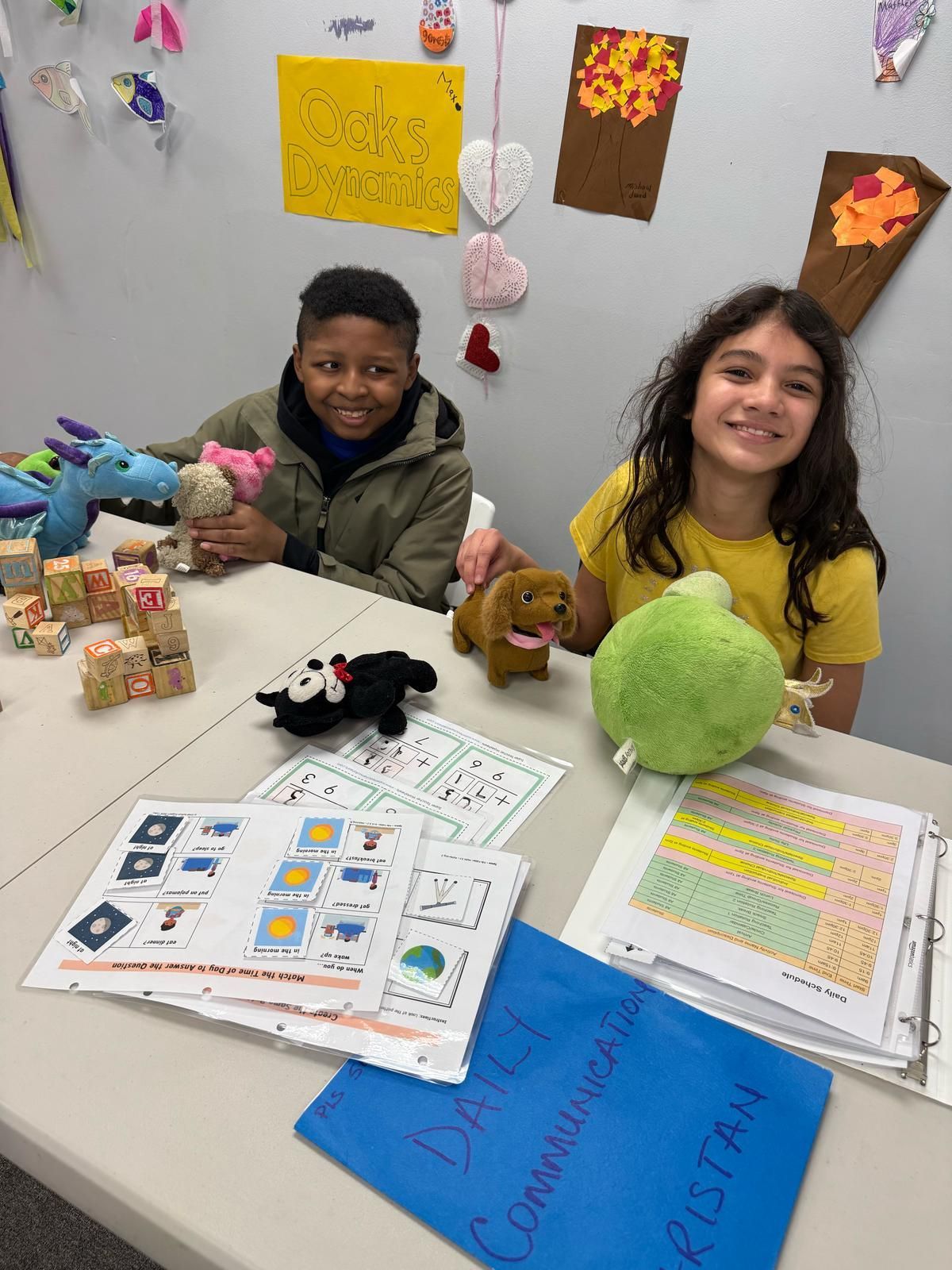 Two children smiling, holding stuffed animals at a crafting table with papers and colorful decorations.