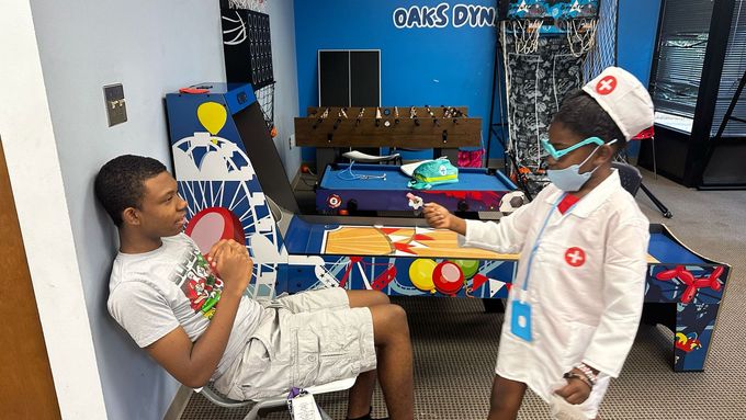 Child in a nurse costume pretends to treat a person seated in a room with games.